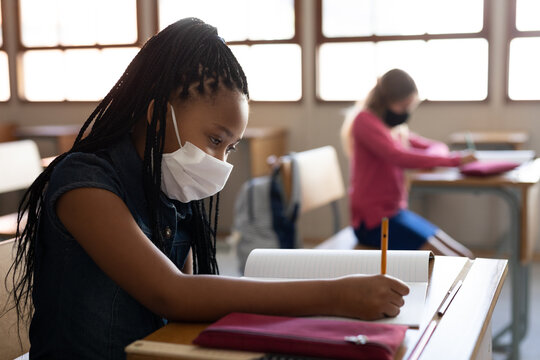 Mixed Race Girl wearing face mask writing while sitting on her desk at school 