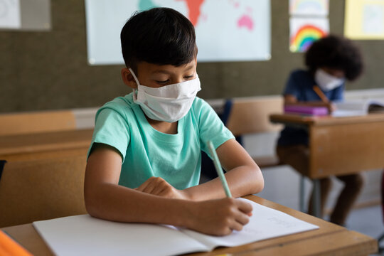Boy wearing face mask writing while sitting on his desk at school 