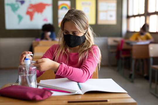 Girl wearing face mask sanitizing her hands while sitting on her desk at school