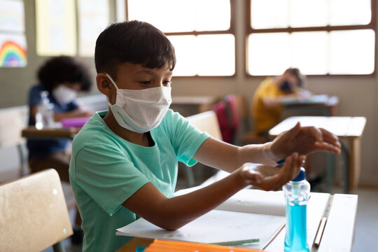 Boy wearing face mask sanitizing his hands while sitting on his desk at school