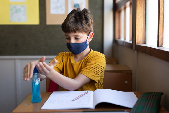 Boy Wearing Face Mask Sanitizing His Hands While Sitting On His Desk At School