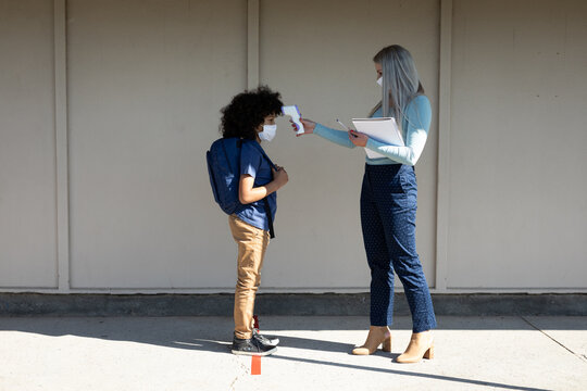 Female teacher measuring temperature of a boy wearing face mask at school