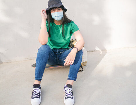 Young beautiful asian woman sitting on a skateboard and wearing protective face mask for coronavirus