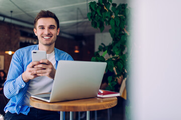 Portrait of happy skilled man connecting to 4g wireless on laptop and mobile technology while posing in coworking space, young Caucasian freelancer working remotely with ideas for blog website