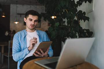 Handsome Caucasian copywriter in smart casual clothing sitting at table with modern laptop and...