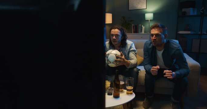 Tensed worried Caucasian male football fans friends sitting in front of TV screen in dark living room on vouch and watching match. Scoring goal or winning game concept. Cheerful men celebrating win.