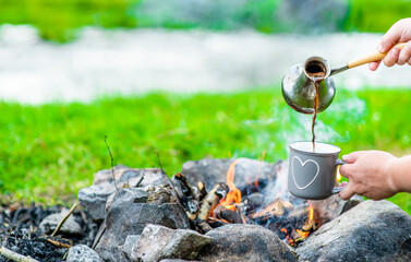 Woman pours hot coffee from a turkish cezve near campfire. Empty space for text