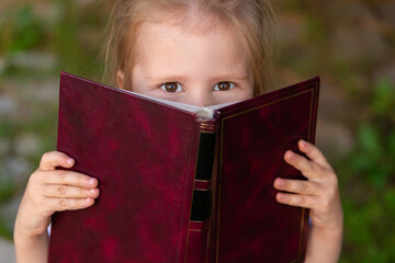 the child slyly narrowed his eyes and holds a book. Little girl hiding behind a book and looking at...