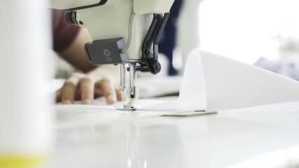 Female hands sewing white sewing machine, close up shot. Female fingers sewing white cloth on white table, selective focus, bright modern atelier studio