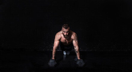 Naklejka premium Young sweaty strong fit muscular man with big muscles doing push ups on the dumbbells in the gym as hardcore cross workout training with free copy space
