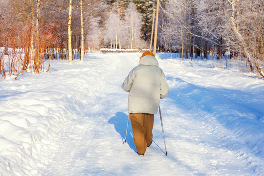 Adult woman in winter sportswear with sticks for Nordic walking on winter landscape city park background, back view. Mature woman doing exercise outdoors. Winter soprt healthy lifestyle concept