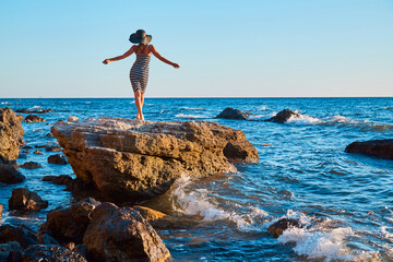 Woman in dress dancing on rock. Waves crashing on stones. Virgin nature, summer holiday and beach concept. Copy space