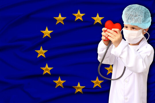 child, boy, in a white doctor’s coat, hat and mask attached a stethoscope to a red heart model, background flag of European Union, close-up, focus on the face, medical concept, cardiology, copy space