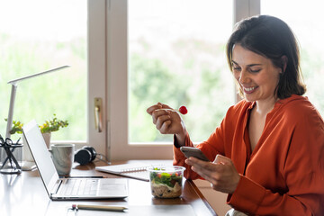 Smiling business person using mobile phone while eating salad at home