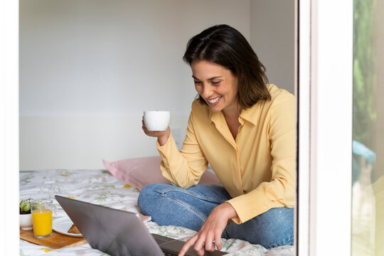 Smiling woman drinking coffee while using laptop in bedroom