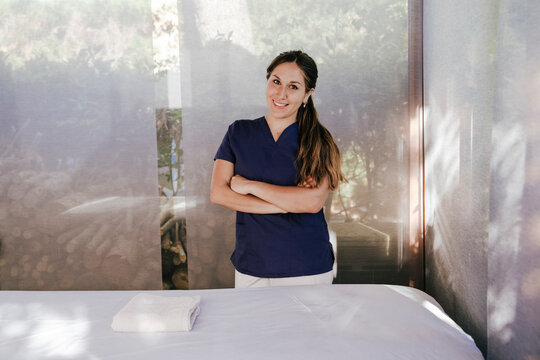 Smiling female massage therapist with arms crossed standing by table in spa