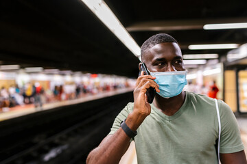 Young man wearing mask talking over smart phone while standing at subway station