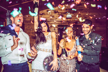 Male and female friends with champagne and disco ball dancing amidst confetti in party