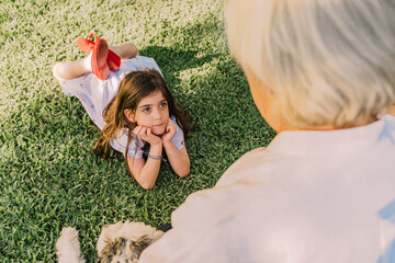 Cute girl with hands on chin looking at grandmother while lying over grassy land in yard