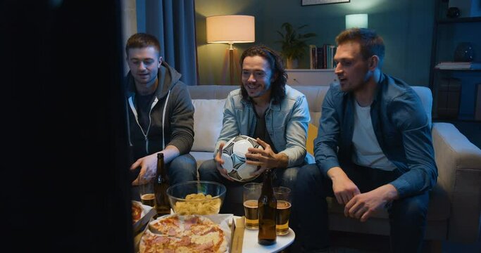 Three Caucasian cheerful best friends eating pizza and drinking beer. Guys sitting on couch at home in evening in front of TV. Football fans watching game on sport channel.