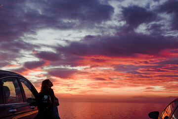 Mid adult woman standing by car at beach against cloudy sky during sunset