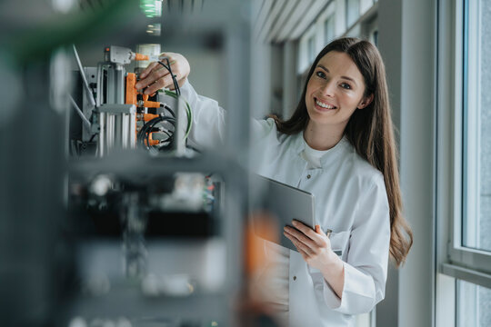 Smiling female scientist holding digital tablet inventing machinery in factory