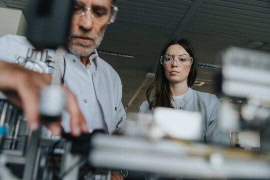 Male and female scientists with protective eyewear examining machine in laboratory