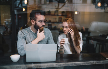 Confused young woman looking at laptop and talking on mobile phone while sitting with boyfriend in cafe
