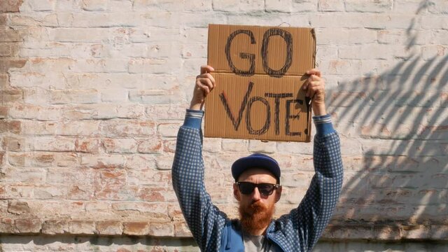Man shows cardboard with Go Vote sign on brick wall urban background. Voting concept. Make the political choice, use your voice. Bearded guy in sunglasses invite to go to the presidential elections.