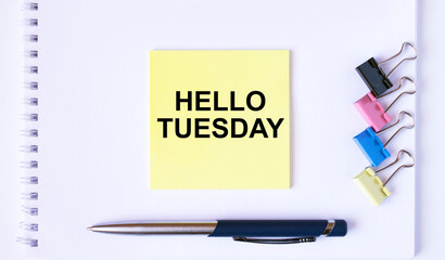 Yellow sticker with text Hello Tuesday lying on a white Notepad with a pen and paper clips