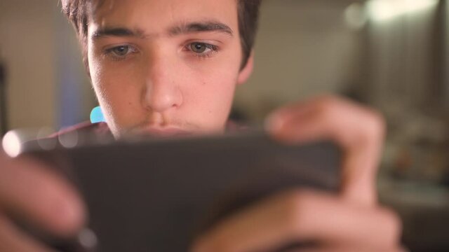 Close up face of a teenage boy with smartphone, at home. Caucasian teen guy spending time in a social network using mobile phone. White young man using cell phone for gaming.