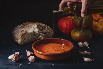 Still life of Salmorejo in a clay plate accompanied by the ingredients with which it is prepared: bread, tomato, garlic and olive oil. Darkfood style photography with matte finishes.