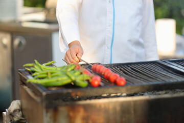 Cook in white coat grill vegetables in summer kitchen. Man hand turn tomato skewer.