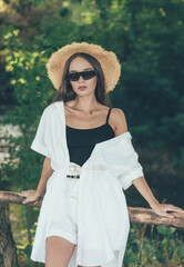 
Stylish slender woman of model appearance in a hat and glasses dressed in a white suit sits in nature near the river