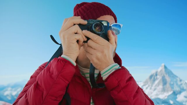 Portrait of millennial travel blogger or social media influencer, hipster man with tattoos in red puffy jacket and beanie hat make photos on analog film camera at mountain top. Wanderlust winter trip