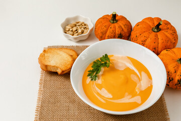 A bowl of pumpkin or carrot vegetarian cream soup is garnished with fresh parsley and cream on a white textured table. selective focus