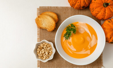 A bowl of pumpkin or carrot vegetarian cream soup is garnished with fresh parsley and cream on a white textured table. flat lay, copy space