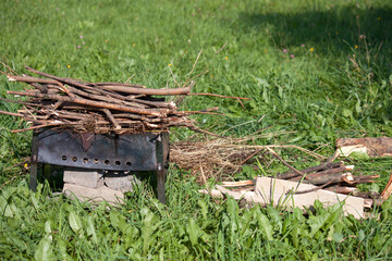 Metal grill with firewood among the green grass in the meadow. Near firewood and grass for lighting.