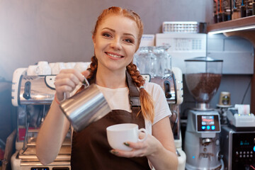Young smiling lady barista in brown apron preparing and coffee order while standing at cafe counter...