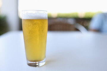 Glass is on white table outside. Glass of yellow cold beer with white foam is poured.