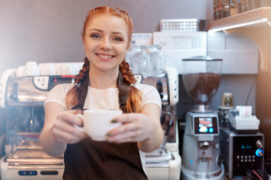 Smiling Cafe Waitress Offering Up Cup Of Fresh Coffee, Red Haired Barista Wearing White T Shirt And Brown Apron Looking Smiling At Camera, Posing Wit Coffee Machine On Background.