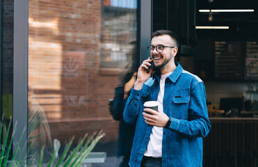 Cheerful male student talking on smartphone while standing with coffee near glass door of coffee shop