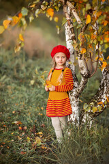 little girl child in a red riding hood is standing by a tree. Autumn leaves around. A girl near a birch tree.