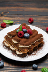 portion of Classic tiramisu dessert with raspberries and blueberries on wooden background