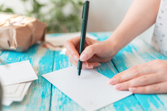 A woman writes on an empty postcard. Hands holding a pen close-up. Blue wooden table with a parcel in the background. The concept of mail correspondence and postcrossing