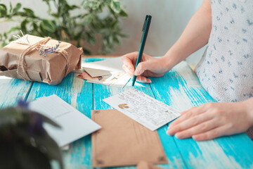 A woman writes a letter, next to it is a craft envelope. Hands lose-up. Blue wooden table with a...