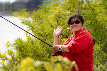 A smiling Asian middle-aged woman, in her 50s, holds a fishing rod and makes a v-sign. Green bushes in the background