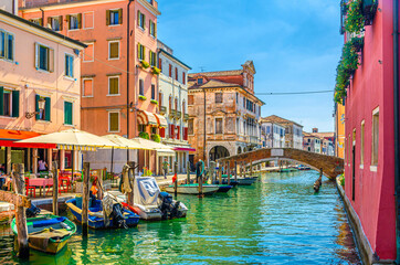 Fototapeta na wymiar Chioggia cityscape with narrow water canal Vena, moored multicolored boats, street restaurant on embankment, old colorful buildings and brick bridge, blue sky in summer day, Veneto Region, Italy