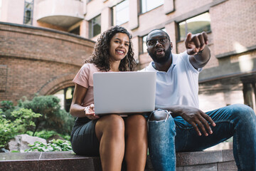 Positive dark skinned male and female millennial students talking to each other using technology outdoors watching video together,african american man pointing and show smth to girlfriend on street