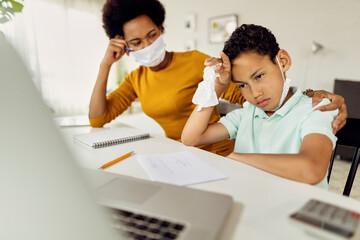 African American boy feeling stressed out while homeschooling with mother during coronavirus...